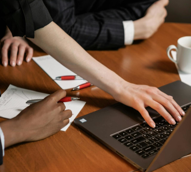 Stół z notatkami i otwartym laptopem, a wokół niego elegancko ubrane osoby. Jedna z nich pisze coś na laptopie, a pozostali notują na kartkach. Zbliżenie na stół, ręce i laptopa